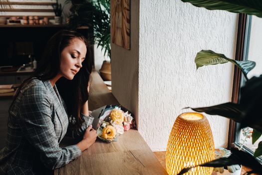 Portrait of a young European girl with long hair in a cafe with a bouquet standing near the window, a tall girl in a jacket with long hair in a cafe waiting.