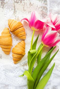 Three croissants and bright pink tulips on lace tablecloth.