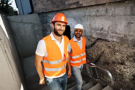 Two young male engineers in uniform and hardhats working at construction site, close up