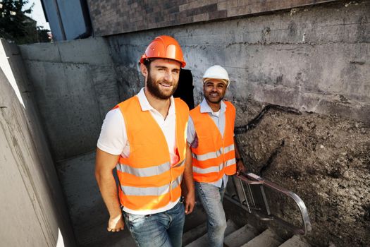 Two young male engineers in uniform and hardhats working at construction site, close up