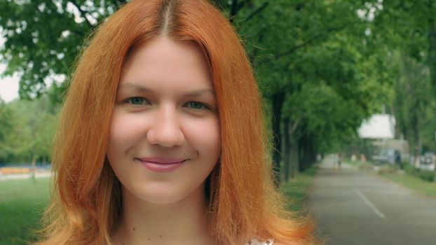 Portrait young cheerful girl with red long hair outdoors in city park. Attractive caucasian redhead woman looking at the camera.