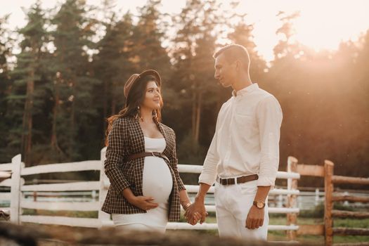 a pregnant girl in a hat and her husband in white clothes stand next to a horse corral at sunset.a stylish couple is waiting for a child in nature.