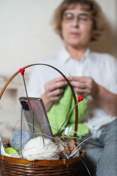 Faceless old woman knits and watches online training on a smartphone. Close-up of female hands with yarn and knitting needles