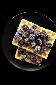 Belgian wafers with frozen blackberries on a black background, close-up.