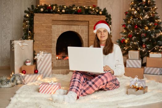 Young woman spending Christmas Eve in cozy christmas interior while sitting on floor and looking directly at camera, wearing casual home attire and santa hat, posing near fir tree and fireplace.