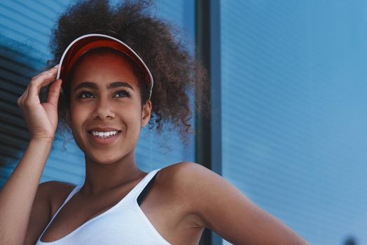 Young woman wearing tennis visor free style on the street walking near window touching hat looking aside smiling cheerful close-up
