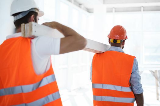 Two young men builders carrying wood planks on construction site, close up photo