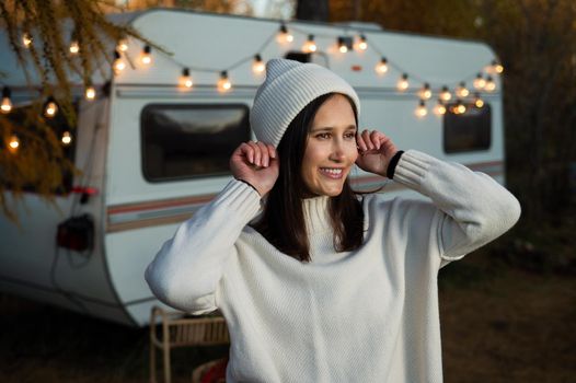 A pensive Caucasian woman in a white knitted sweater and a hat is resting near a motorhome in a warm autumn. Travel by camper