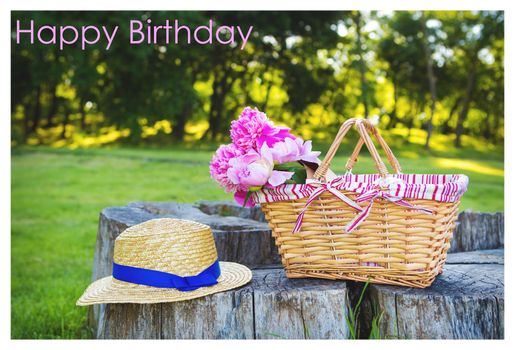Beautiful straw hat and flowers in a basket stand on a wooden stump-happy birthday inscription