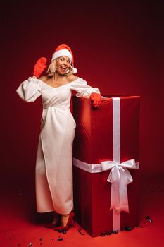 portrait of a smiling girl in a white dress and Santa hat with a huge Christmas gift on a red background.