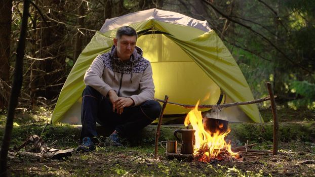 The man sits near the fire. Camping in a beautiful forest. The food in the cauldron is cooked over the fire. Travel concept