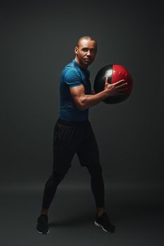 Full length shot of a crossfit male athlete working out with exercise ball. Handsome muscular athletic sportsman exercising with exercise ball over dark background in studio. Endurance concept