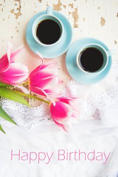 two cups of coffee and pink tulips on an old table, writing happy birthday.