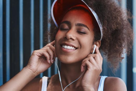 Young woman wearing tennis visor and earphones free style on the street standing near fence head up closed eyes smiling delightful close-up