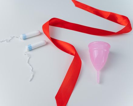 Tampon, pink menstrual cup and red satin ribbon on a white background. Means of intimate hygiene of a woman during mentruation.