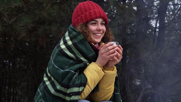 Young woman drinks tea by the fire in the winter forest. She is smiling and happy. Travel and adventure concept.