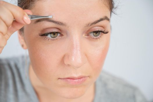 Close-up portrait of a caucasian woman doing eyebrow correction herself with tweezers.