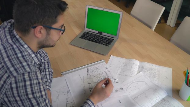 Young professional designer checking architectural sketch at the working place with laptop pencil line equipment for drawing and draftsmanship. On the big desk plan new building or house guy draw some details. Caucasian mixed race male wearing in casual checkered blue shirt with glasses.