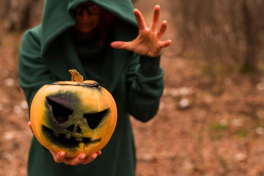 A creepy witch holds a steaming pumpkin in a deep forest. Jack o lantern emits yellow smoke for halloween.