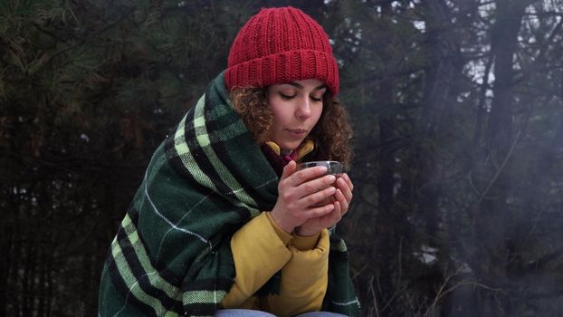 Young woman drinks tea by the fire in the winter forest. She is smiling and happy. Travel and adventure concept.