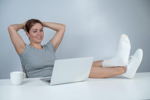 Caucasian woman lifted her leg with plaster to work desk and works on laptop on white background.