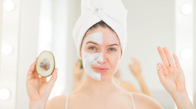 A woman with a towel on her hair and in a clay face mask holds an avocado. Taking care of beauty at home.