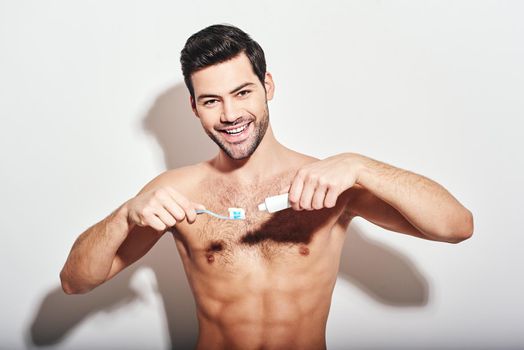 Handsome young man squeezes the toothpaste on a toothbrush while standing over white background and smiling at camera