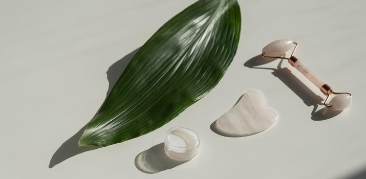 Pink gouache scraper, roller, jar of green leaf cream on a white table. Equipment for self-massage and skin care for the face and neck.