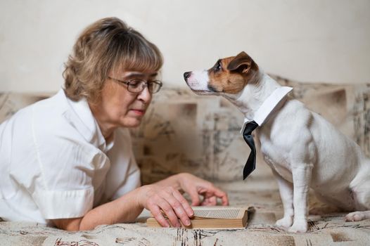 An elderly caucasian woman is lying on a sofa with a smart dog jack russell terrier wearing glasses and a tie and reading a book