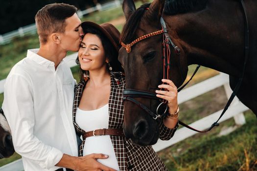 a pregnant girl in a hat and a man in white clothes stand next to horses near a white fence.Stylish pregnant woman with a man with horses.Married couple