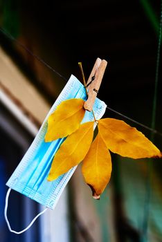 A blue medical mask hangs on a rope along with a branch of brown leaves. Autumn mask mode concept