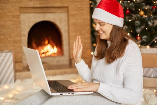 Beautiful young woman in santa hat greets Christmas or New Year online while sitting by laptop on her knees against background of Christmas tree and fireplace, waving hand to camera.