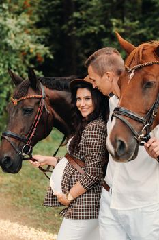 a pregnant girl in a hat and her husband in white clothes stand next to horses in the forest in nature.Stylish pregnant woman with a man with horses.Family
