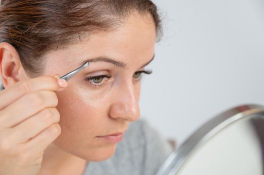 Caucasian woman looks in the mirror and does the eyebrow correction herself with tweezers.