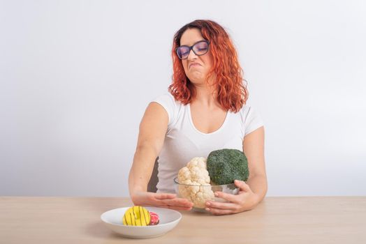 Caucasian woman prefers healthy food and refuses fast food. Redhead girl chooses between broccoli and donuts on white background.