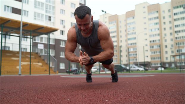 Young muscular man doing push up exercise on the ground outdoors. He looking at camera. The guy presses on the sports ground on the street.