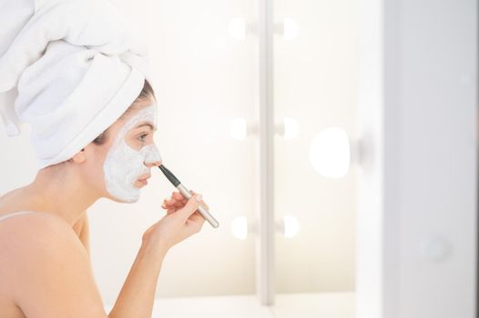 Cheerful woman with a towel on her hair and in a clay face mask fooling around with cucumbers in her hands. Taking care of beauty at home.