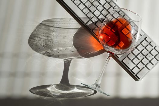 A glass of red wine lies on the keyboard on a white table with a shade from the blinds.