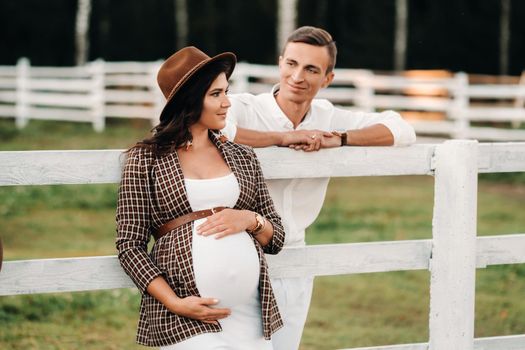 a pregnant girl in a hat and her husband in white clothes stand next to a horse corral at sunset.a stylish couple is waiting for a child in nature.