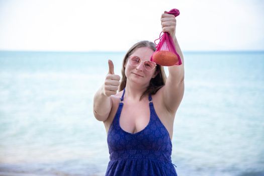 woman holding mesh bag with fruit and showing thumbs up. on the background is sea or ocean.
