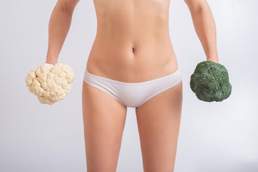 A faceless woman in panties holds cauliflower and broccoli on a white background. Food habits