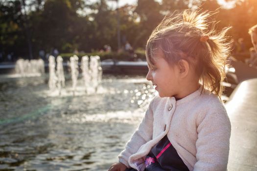 adorable toddler girl looks at the pond with fountains in the park on a sunny day. weekend family walk. spending time with children. artistic focus