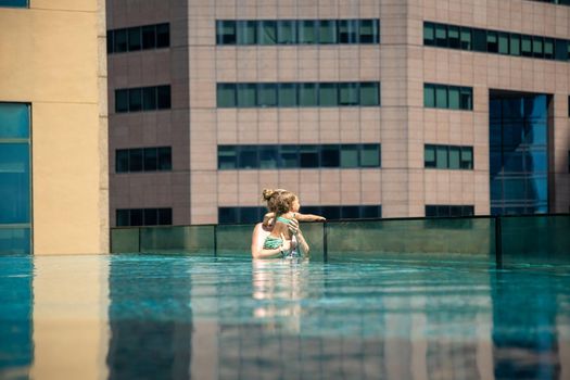 young woman with her little daughter in the pool on the roof of a skyscraper in a metropolis look overboard...