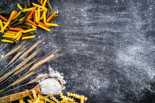 spikelets of wheat, pasta and spoon with flour on a black textured table