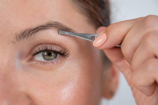 Close-up portrait of a caucasian woman doing eyebrow correction herself with tweezers.