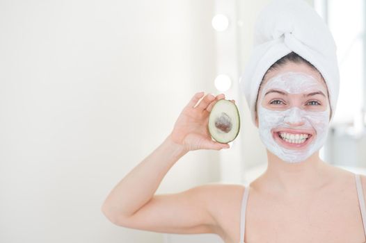 A woman with a towel on her hair and in a clay face mask holds an avocado. Taking care of beauty at home.