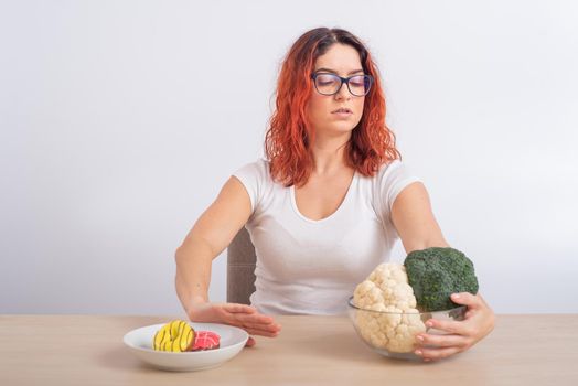 Caucasian woman prefers healthy food and refuses fast food. Redhead girl chooses between broccoli and donuts on white background.
