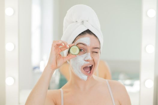 Cheerful woman with a towel on her hair and in a clay face mask fooling around with cucumbers in her hands. Taking care of beauty at home.