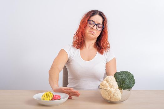 Caucasian woman prefers healthy food and refuses fast food. Redhead girl chooses between broccoli and donuts on white background.