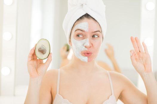 A woman with a towel on her hair and in a clay face mask holds an avocado. Taking care of beauty at home.